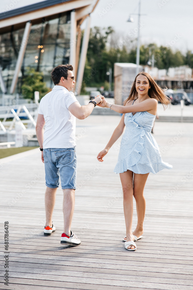 Fototapeta premium A couple in love, a man and a woman, walk along a wooden pier. A man and a woman hold hands and run along a wooden pier. Stylish man and his beautiful woman smiling and having fun