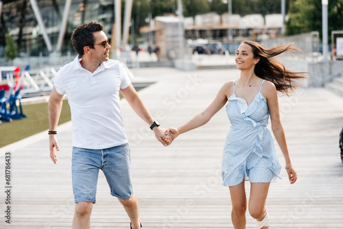 A couple in love, a man and a woman, walk along a wooden pier. A man and a woman hold hands and run along a wooden pier. Stylish man and his beautiful woman smiling and having fun