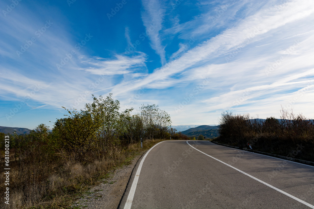 Asphalt road a top a mountain, framed by a vivid blue sky streaked with picturesque white clouds. The empty, newly laid two lane road leads toward distant hills and mountains