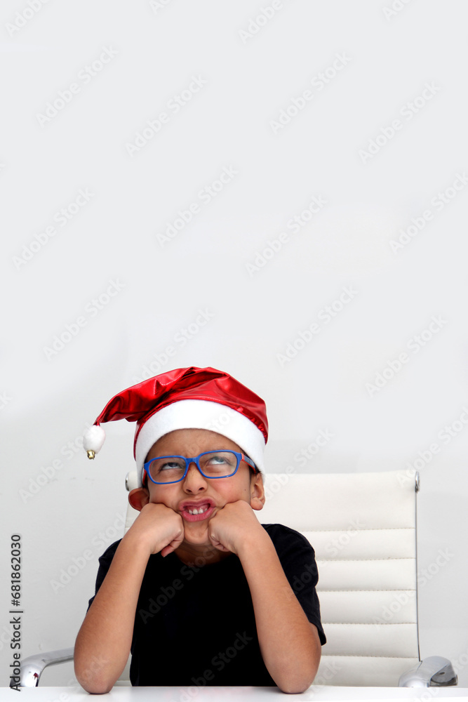 Obraz premium 7-year-old dark-skinned Latino boy with a Santa Claus Christmas hat looks sad and angry because he doesn't like the holidays and they didn't give him what he wanted