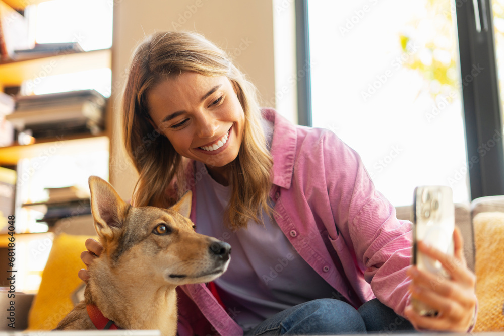 Smiling beautiful woman, influencer holding mobile phone taking selfie with cute dog, recording video sitting in modern cafe  - obrazy, fototapety, plakaty 