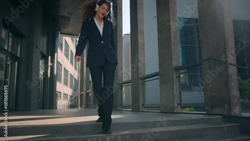 Happy young business woman in formal suit dancing walking outdoors listening music headphones audio joyful funny female Caucasian businesswoman girl go down steps of building dance in city sunlight  photo