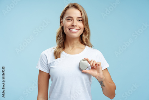 Portrait of attractive woman in white t shirt holding pacemaker in hand, looking at camera isolated on blue background. Female with silky wavy hair standing in studio. Health care, treatment concept photo