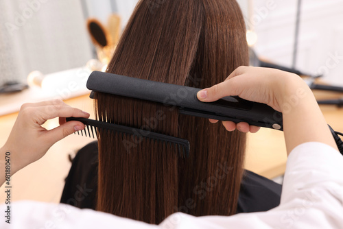 Hairdresser straightening woman's hair with flat iron in salon, closeup
