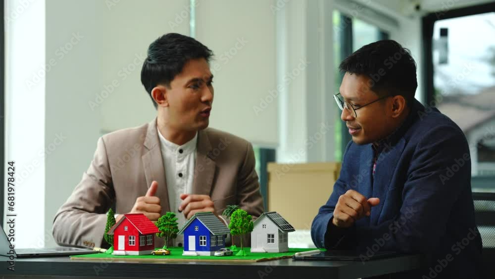 Two professionals smiling, discussing over model houses on a table, possibly planning or reviewing a housing project on large land. Asian people, only businessman, Middle-age Two professionals smiling, discussing over model houses on a table, possibly planning or reviewing a housing project on large land. Asian people, only businessman, Middle-age