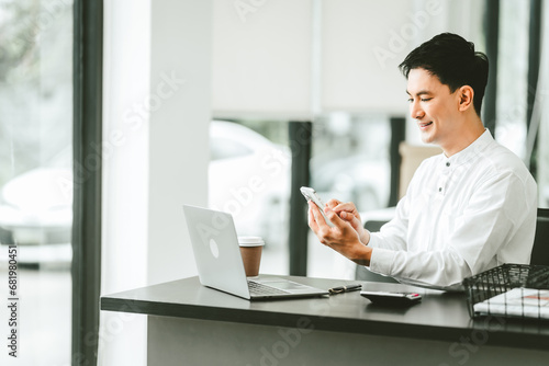 Attractive smiling male asian business person in white shirt working on laptop at desk with coffee cup and documents nearby.