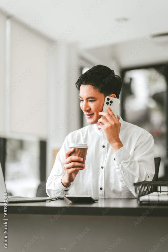 Attractive smiling male asian business person in white shirt working on laptop at desk with coffee cup and documents nearby. Attractive smiling male asian business person in white shirt working on laptop at desk with coffee cup and documents nearby.