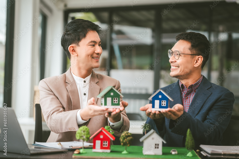 Two professionals smiling, discussing over model houses on a table, possibly planning or reviewing a housing project on large land. Asian people, only businessman, Middle-age