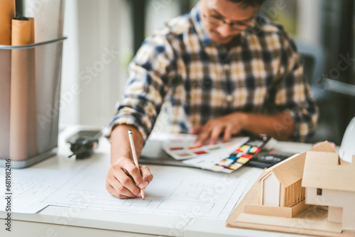 Asian businessman person with glasses examining architectural blueprints next to model house, engineer architect working on new house project and apartment.