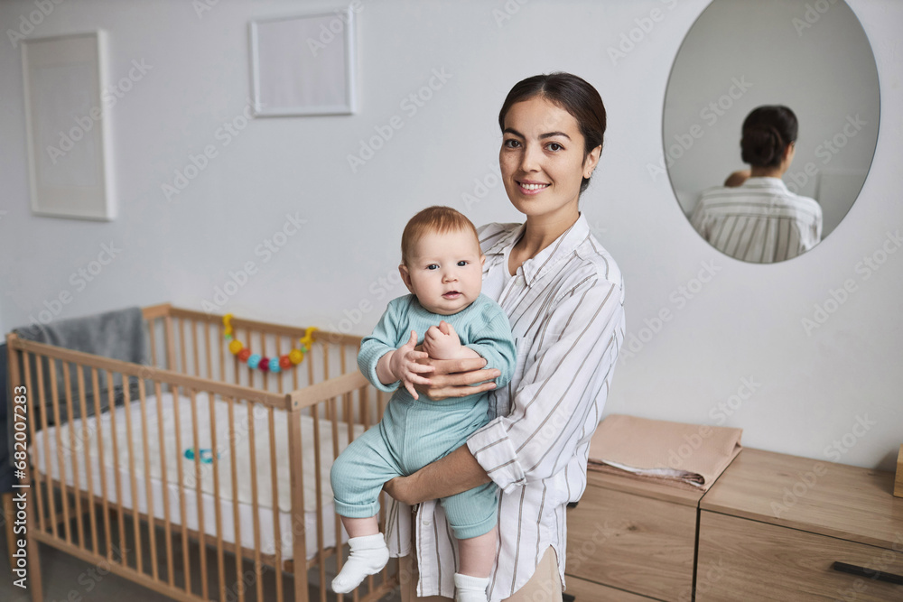 Waist up portrait of happy young mother holding baby son in cozy room, both looking at camera Waist up portrait of happy young mother holding baby son in cozy room, both looking at camera