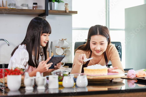 Asian little girl baker pastry bakery chef daughter wear apron standing holding tablet computer learning online when female mother smiling using whipped cream decorating homemade cake in home kitchen