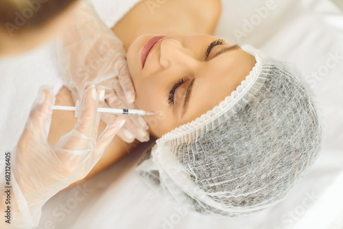 Beauty specialist injects neurotoxin or dermal filler in crows feet or upper eyelid. Close up woman's head in white cap and doctor's hands in gloves. Aesthetic face skin eye wrinkle treatment concept photo