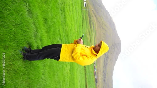 Man wearing yellow raincoat using phone in Faroese Vidareidi's grass field. Vertical photo