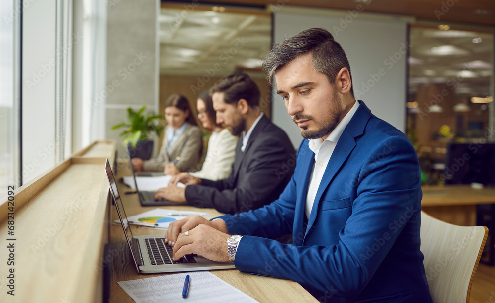 Group of busy people working in modern coworking space or business center. Man in formal suit sitting at desk by window with other men and women, studying data reports and working on laptop computer Group of busy people working in modern coworking space or business center. Man in formal suit sitting at desk by window with other men and women, studying data reports and working on laptop computer
