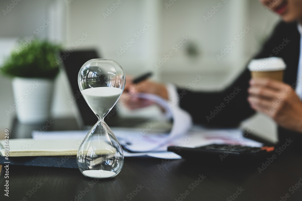 Closeup hourglass on office desk with blurred businessman working on background Closeup hourglass on office desk with blurred businessman working on background