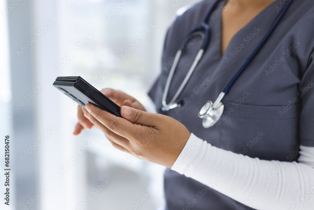 Midsection of biracial female doctor wearing scrubs using smartphon in hospital