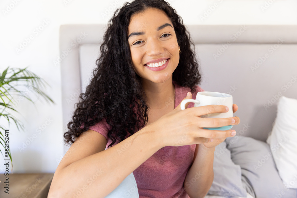 Portrait of happy biracial woman holding cup of coffee sitting on bed at sunny home Portrait of happy biracial woman holding cup of coffee sitting on bed at sunny home