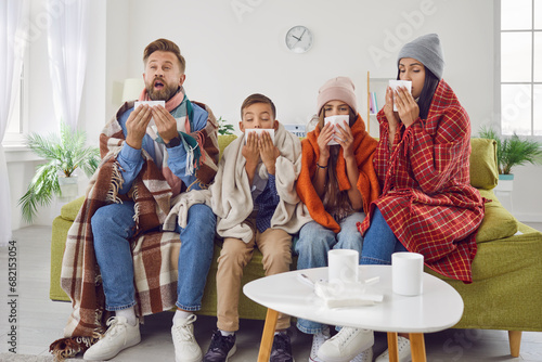 Achoo. Family of four have caught bad cold all together. Sick mom, dad and children in warm clothes and plaids sitting on couch in living room and sneezing in paper handkerchiefs. Winter, flu concept photo