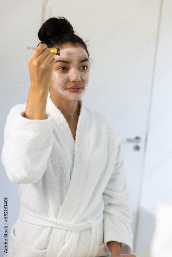 Biracial woman in bathrobe applying face mask using brush in sunny bathroom Biracial woman in bathrobe applying face mask using brush in sunny bathroom