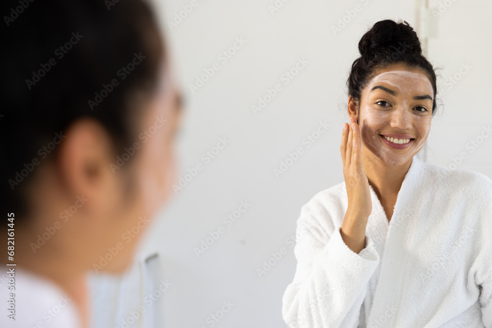 Happy biracial woman in bathrobe with face mask in sunny bathroom Happy biracial woman in bathrobe with face mask in sunny bathroom