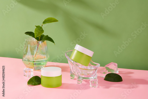 Two jars of lotion with green tea extract are displayed with fresh green tea leaves and glass props on a green-pink background. Blank label for design or beauty product promotion.