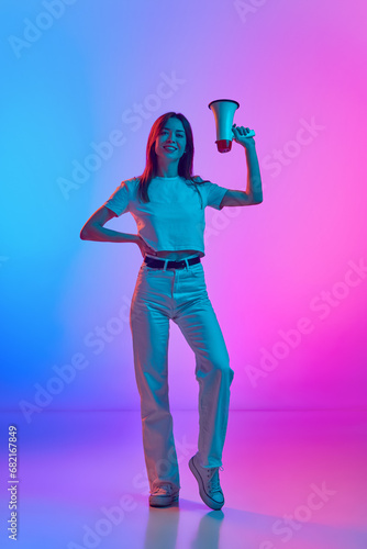 Full-length image of elegant woman in her 30s standing with loudspeaker over gradient pink blue studio background in neon light. Concept of human emotions, lifestyle, youth culture, facial expression