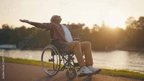 Senior man in wheelchair enjoys freedom. Male with special need spreading arms on sides smiling breathing fresh air walking in city park near pond. Living with physical disability in old age concept. photo