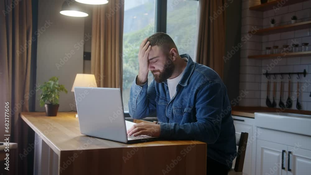 Bearded young adult man sitting against the kitchen counter using laptop having anxiety and stress. Upset man reacting to loss, bad news Bearded young adult man sitting against the kitchen counter using laptop having anxiety and stress. Upset man reacting to loss, bad news