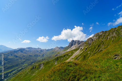 Eine weite Berglandschaft mit saftig grünen Hängen erstreckt sich unter einem strahlend blauen Himmel mit vereinzelten weißen Wolken.