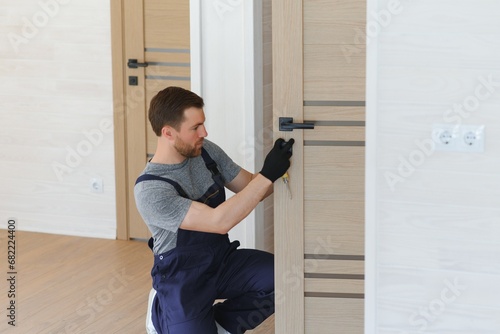 Installation of a lock on the front wooden entrance door. Portrait of young locksmith workman in blue uniform installing door knob. Professional repair service. Maintenance Concept © Serhii