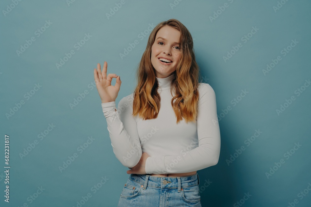 Smiling woman making OK sign with hand, dressed in white turtleneck and jeans on a blue background. Smiling woman making OK sign with hand, dressed in white turtleneck and jeans on a blue background.