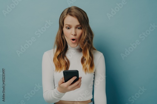 Shocked young woman holding phone, mouth agape, expressing surprise, dressed in white on a teal background