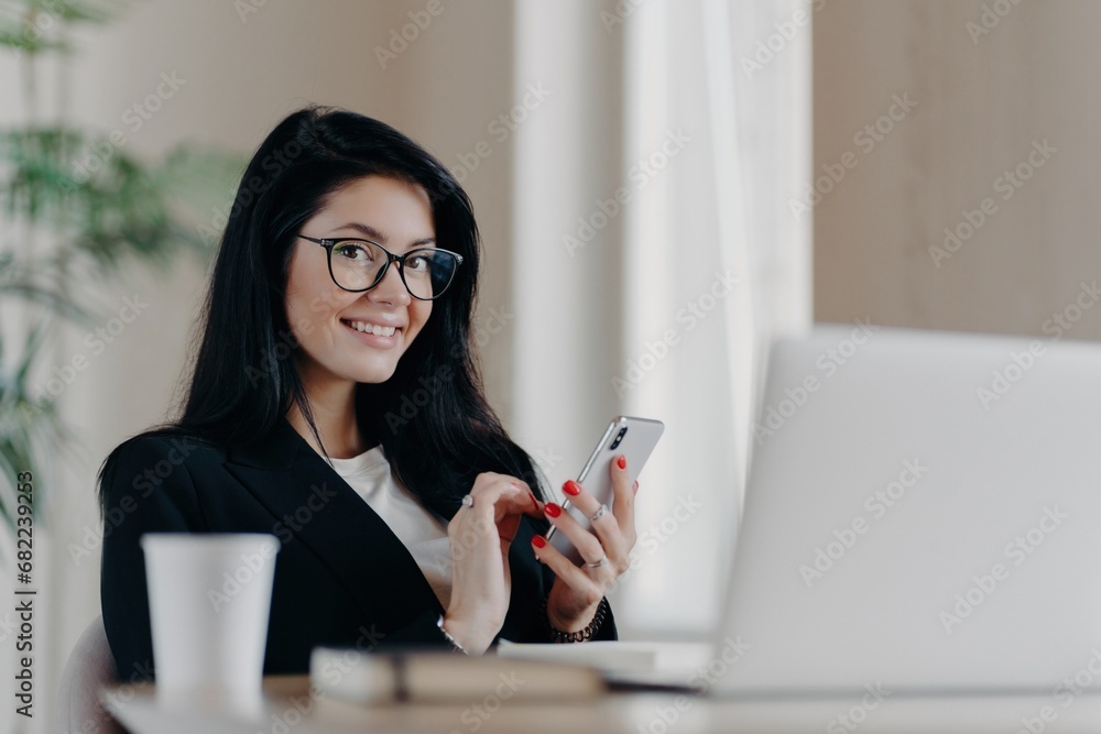 Smiling businesswoman using smartphone at her workspace with laptop and coffee cup in background Smiling businesswoman using smartphone at her workspace with laptop and coffee cup in background