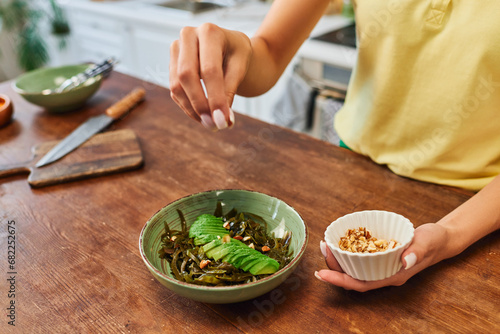 cropped woman adding walnuts into vegetarian salad with seaweed and sliced avocado, plant-based diet