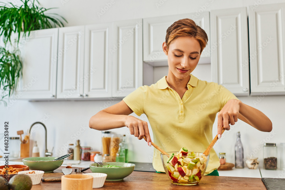 young smiling woman mixing delicious fruit salad while cooking in kitchen, plant-based diet concept