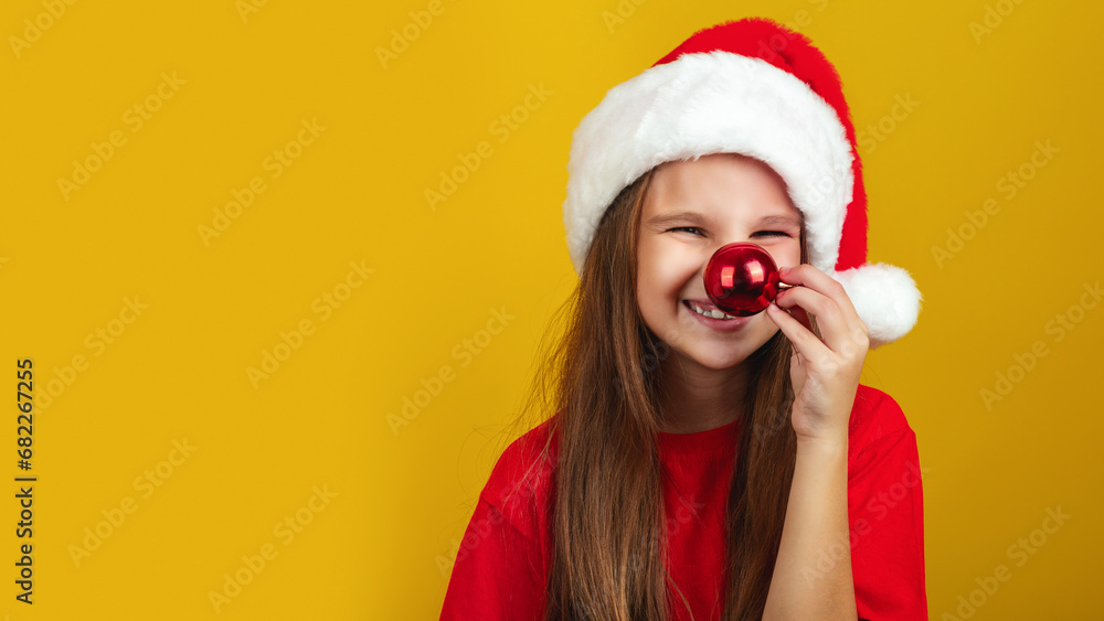 Holiday Fun. Cheerful Little Girl Wearing Santa hat, Playful Kid Keeping Xmas Tree Ball Near Nose And Looking At Camera, Yellow Background. Copy Space Holiday Fun. Cheerful Little Girl Wearing Santa hat, Playful Kid Keeping Xmas Tree Ball Near Nose And Looking At Camera, Yellow Background. Copy Space