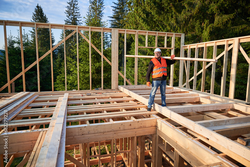 Carpenter builds wooden frame house near woods. Man holds large beam on shoulder, wearing work clothes and helmet. Concept of modern and sustainable building.
