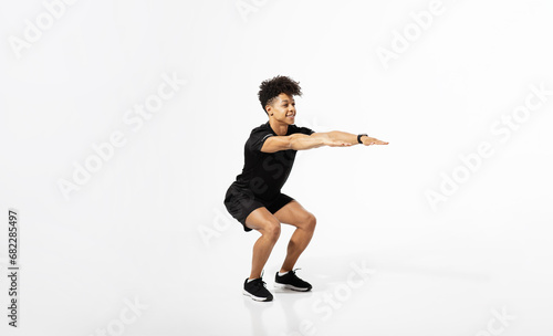 Athletic young man extending arms squatting over white studio background