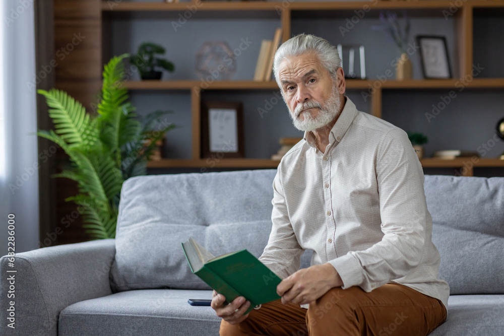 Portrait of a gray-haired senior man sitting at home on the couch, holding a book in his hands and looking seriously into the camera Portrait of a gray-haired senior man sitting at home on the couch, holding a book in his hands and looking seriously into the camera