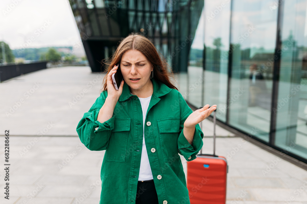 Unhappy 30s woman tourist dressed in casual look speaking on smartphone gadget on the city street. Angry redhead girl a red suitcase standing on city street. Negative people emotion.