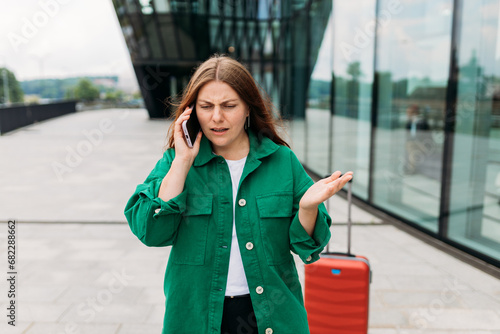 Obraz na płótnie Unhappy 30s woman tourist dressed in casual look speaking on smartphone gadget on the city street. Angry redhead girl a red suitcase standing on city street. Negative people emotion. w salonie