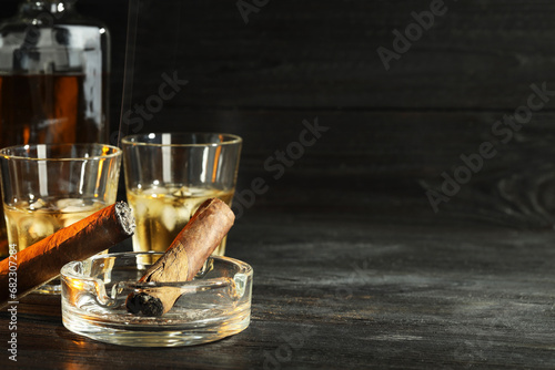 Cigars, ashtray and whiskey with ice cubes on black wooden table. Space for text