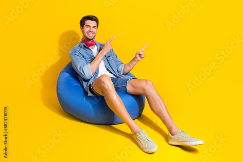 Full size photo of positive guy wear jeans jacket shorts indicating at logo empty space sit on pouf isolated on yellow color background