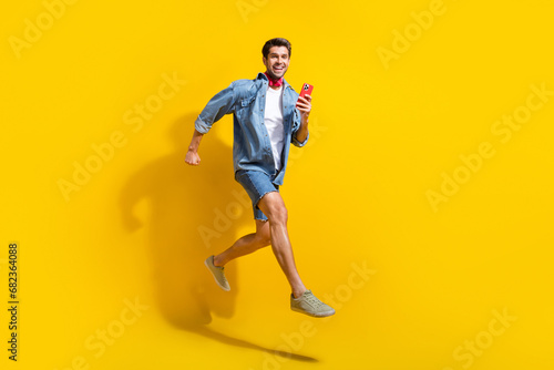 Full size photo of excited positive guy wear jeans jacket scarf run shopping holding smartphone in arm isolated on yellow color background