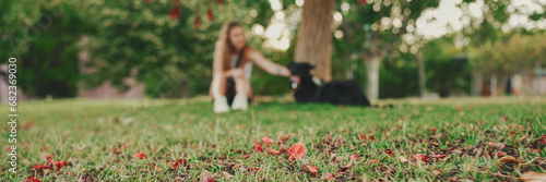 Cute girl with black dog is resting under tree on the lawn. Happy dog rejoices being on walk in the outdoor. Animal training outdoors.