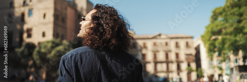 Young italian guy with long curly hair and stubble walks along the fenced sidewalk and looks around.