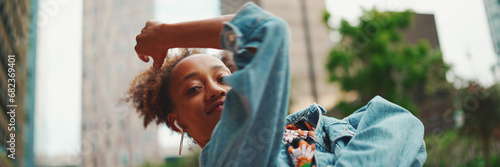 Closeup, smiling African girl with ponytail wearing denim jacket, in crop top with national pattern listening to music on headphones and dancing outdoors. photo