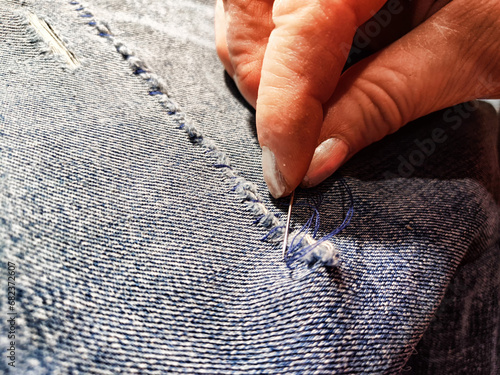 Woman repairing a pair of blue jeans. Fingers with a needle and torn jeans fabric. Sewing up a tear on a blue fabric