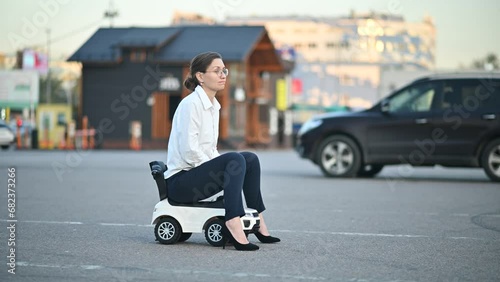 A black sedan leaves the parking lot and we see a young woman in a toy car sadly starting to drive a children's car in the parking lot.