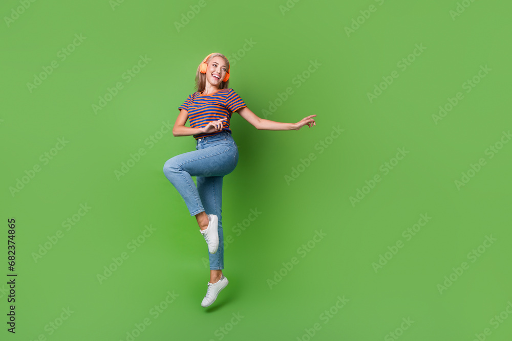 Full length photo of youngster girl jumping music wireless earphones dancing at home atmosphere party isolated on green color background